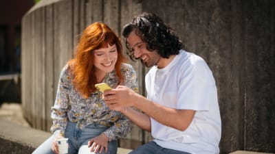 En ung kvinna och en man sitter brevid varandra och ser på något på mannens telefon. 