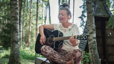 Nainen istuu penkillä risti-istunnassa, soittaa kitaraa ja laulaa, taustalla keväistä metsää