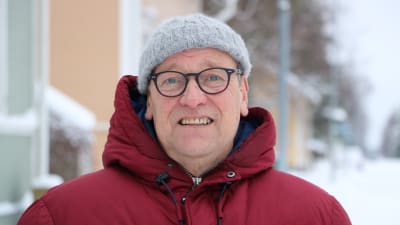 En man glasögon, grå mössa på huvudet och en röd dunjacka med huva, står på en vinterklädd gata.