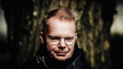 En man i runda glasögon och svart jacka står framför en trädstam.