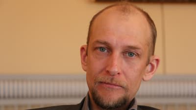 En man med kort hårt och skägg sitter i en fullmäktigesal.