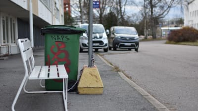 En busshållplats vid en gata i Hangö, i förgrunden en vit bänk och en grön soptunna där någon klottrat med röd färg och skrivit NAPS med versaler.