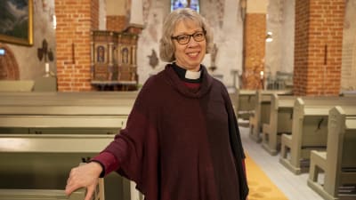 En kaplan ser mot kameran och ler. Hon står i en gammal kyrka.