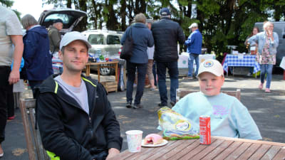 På bilden sitter Gustav och Edvin Westerlund. Gustav har en kopp kaffe och en berlinermunk framför sig, och Edvin har meterlakrits. I bakgrunden ser man torgbesökare. Bilden är tagen på Vessö sommartorg.