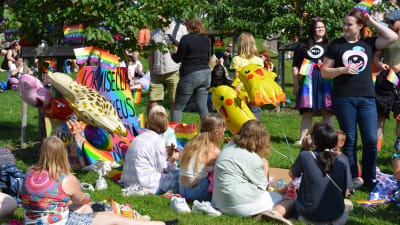 Ungdomar sitter på en gräsmatta och håller picknick med färggranna ballonger och skyltar bredvid sig efter en prideparad.