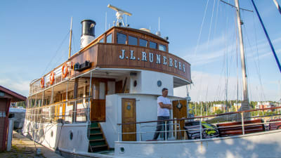 Båt som det står "J. L Runeberg" på förtöjd vid en hamn. En person står på däcket. 