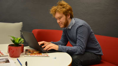 Jakob löfgren, en man med stort och krulligt hår sitter i en röd soffa och skriver på sin bärbara dator.
