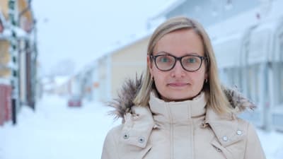 En kvinna med axellångt blont hår och glasögon står på en vinterklädd gata.