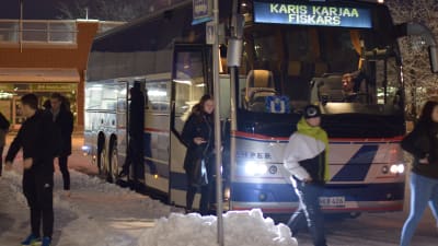 En buss som står stilla vid en busstation. Ungdomar stiger ut ur bussen. Det är vinter.