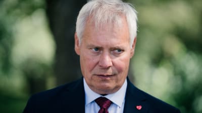 Antti Rinne i mörk kostym och röd slips mot en grön suddig bakgrund. Han har en sammanbiten min.