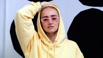 Den svenska hiphop-artisten Silvana Imam sitter med ena handen bakom huvudet, hon har på sig glasögon och en gul munkjacka.