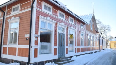 En bild på ett hus som är orangerött och som ligger i gamla stan i Ekenäs. Huset har stora fönster och landskapet runt det är täckt av snö. Huset har en grå dörr.