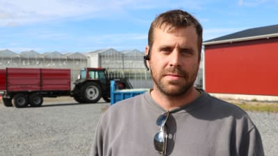 En man med kort brunt hår och skägg står framför ett stort växthus. I bakgrunden syns också en traktor med släp.