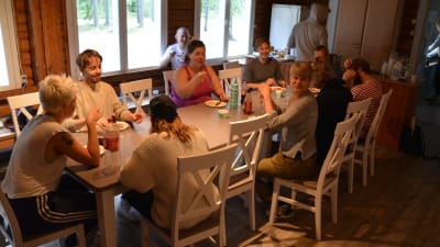 Många glada unga människor sitter kring ett stort matbord i ett kök.