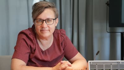 Nina Fellman har röd skjorta, svartbågade glasögon och kort brunt hår. Hon tittar in i kameran med armarna i kors.