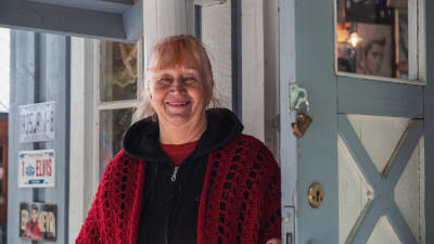 Rödhårig äldre kvinna i svart tröja och röd sjal står i dörröppningen till ett trähus målat i ljusblått.
