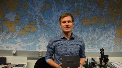 Ung man i blå skjorta håller upp en svart bok och står framför karta över skärgården