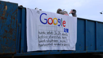 Ett lakan hänger över ett lastbilsflak. På lakanet står det google, samt flera olika sökfraser på finska, som ska se ut som att man gjort olika sökningar på google.