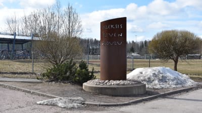 Skylt där det står Aleksis Kiven koulu. Skylten är vid en gräsplan och grusväg.