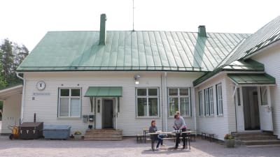 En gammal vit skolbyggnad i trä med grönt koppartak. Framför huset står ett bord där tre kvinnor dukar upp kaffekoppar.