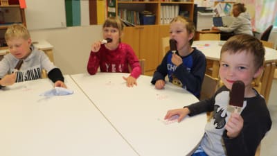 Fyra dagisbarn sitter och äter glasspinnar runt ett bord. En pojke visar upp sin glass för kameran och ler.