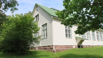 Rödskog skolbyggnad. Ett vitt trähus. Det är sommar och gräset framför huset är grönt och träden brevid huset är fulla med löv.