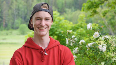 Pojke står och ler med skärmmössan bakvänd vid en rosenbuske.