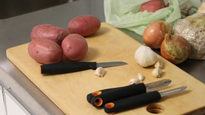 Ett skärbräde med några potatisar, en vitlök och några lökar samt en liten kniv och två skalknivar. 