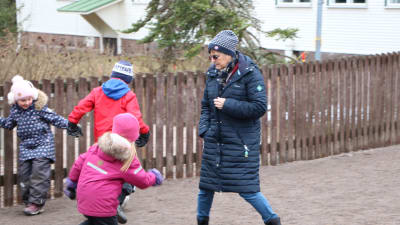 En vuxen kvinna och några barn på en skolgård, iklädda vinterkläder. 