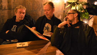 Tre män som pratar med varandra medan de ser på en LP-skiva.