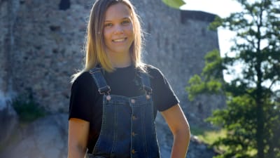 Kvinna i snickarbyxor ler mot kameran. I bakgrunden en medeltida slottsruin.
