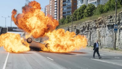 En man står framfölr en bil som exploderat i ett enorms moln av eld.