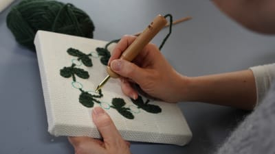 Händer som tuftar fram ett bladmotiv på ett uppspänt tyg