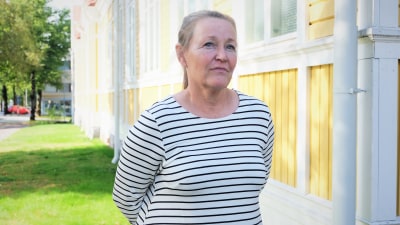 en kvinna i medelålderna med grått hår och randig skjorta som står framför ett gammalt trähus.