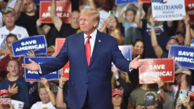 På bilden syns ex-presidenten Donald Trump på ett av sina politiska evenemang. I bakgrunden står hans anhängare med skyltar i blått, rött och vitt där det står "Save America". 