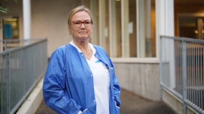 En sjuksötare i vita kläder och knallblå tröja står utanför ingången till jouren vid Raseborgs sjukhus. Höst.
