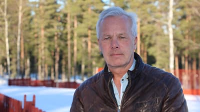 En medelålders man med brun jacka står intill ett rött staket.