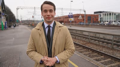 En man i kostym och jacka står vid en perrong på en tågstation. Han heter Henrik Wickström.