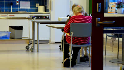 Ett par äldre personer sitter vid ett bord. I bakgrunden syns ett kök.