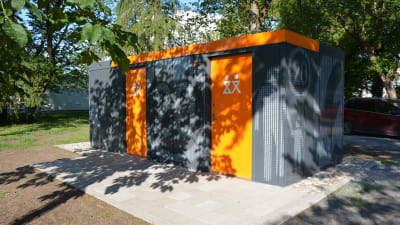 ny offentlig toalett i kuppisparken i åbo. grå rektangylär byggnad med orangea dörrar som stark kontrast.