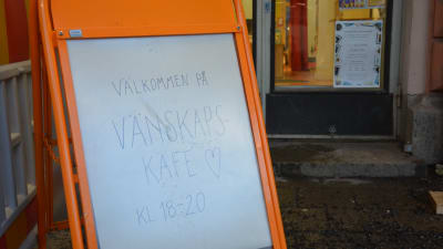 En orange skylt med texten "Välkommen på vänskapskafé kl. 18-20"