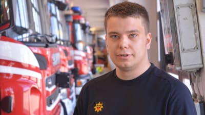 aatu isotalo står i räddningsverkets garage. han bär arbetskläder och i bakgrunden figurerar en rad av brandbilar.