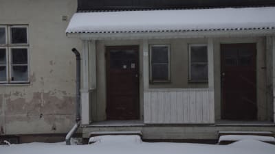 Ett vitrappat hus  där rappningen delvis fallit bort och ett stuprör hänger också löst. Vinter och snö. Huset är obebott.