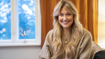 En ung kvinna med långt blont hår och iklädd beige tröja ser in i kameran och ler. Orange gardiner och ett fönster i bakgrunden.