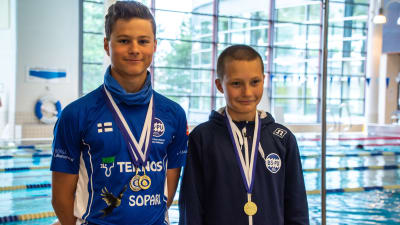 Två unga män eller pojkar står framför en simbassäng i Borgå simhall. De har medaljer runt nacken.