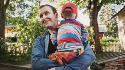 Mies farkkupaita päällään sylissään pieni poika, joka selin kameraan, pojalla raidallinen paita ja punainen lippalakki, taustala vehreää pihaa