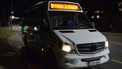 Det är mörkt. Bussen är vit och framtill lyser texten "Byaskjussen".