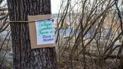 En skylt fäst på ett träd. På skylten har någon på finska skrivit en kort dikt om att den sagolika parken snart kan försvinna.