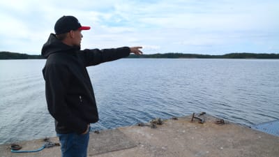En man iklädd svart jacka och keps står och pekar ut på havet