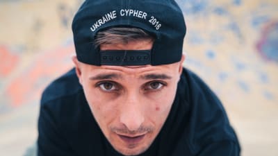 Mies katsoo tiiviissä kuvassa suoraan kameraan. Lippalakki on väärinpäin päässä ja siinä näkyy teksti Ukraine Cypher 2016.